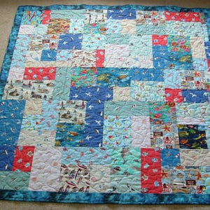 May include: A patchwork quilt with a nautical theme. The quilt features squares of various fabrics in shades of blue, red, and white, with images of sailboats, sea creatures, and beach scenes. The quilt is bordered with a darker blue fabric.
