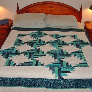 May include: A white quilt with a blue and green geometric pattern. The quilt is on a bed with a wooden headboard and two nightstands.