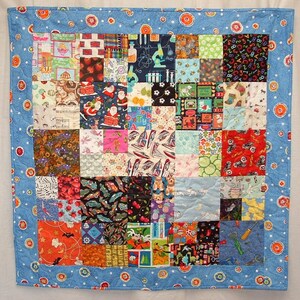 May include: A blue and white patchwork quilt with a variety of colorful squares featuring different patterns and designs. The quilt is surrounded by a blue border with white polka dots.