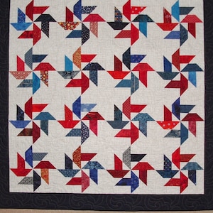 May include: A quilt with a repeating pattern of red, blue, and white pinwheels. The quilt is made of fabric scraps and is quilted with a white thread.
