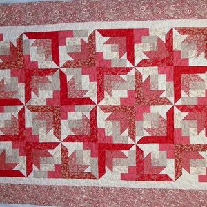 May include: A red, pink, and white quilt with a geometric pattern. The quilt is made of fabric squares sewn together to create a repeating design.