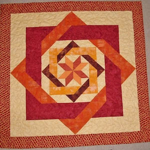 May include: A quilt with a geometric pattern in shades of orange, red, and brown. The quilt is made of fabric squares sewn together to create a starburst design.