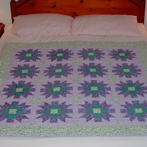 May include: A handmade quilt featuring a repeating starburst pattern in shades of purple and green on a light purple background. The quilt is on a bed with white sheets, and a floral patterned border. The bed is flanked by two wooden nightstands.