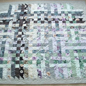 May include: A handmade quilt with a woven design. The quilt features a mix of floral and geometric patterns in shades of gray, white, black, and purple. The edges are finished with a light green border and a quilted pattern.
