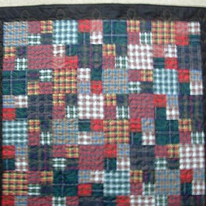 May include: A patchwork quilt with a black border and a variety of plaid patterns in red, green, blue, and white. The quilt is made of squares of fabric stitched together, creating a textured surface. The border has a decorative pattern.
