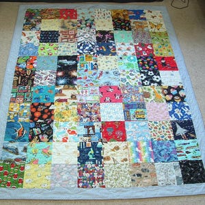 May include: A patchwork quilt with a pale blue border. The quilt is composed of many fabric squares, each with a unique pattern, such as cartoon characters, animals, and other designs. The quilt is rectangular and lies flat on a surface.