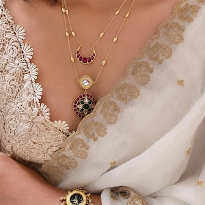 May include: A gold-toned layered necklace featuring a crescent-shaped pendant with red stones and a circular pendant with a central green stone surrounded by red and clear accents. A matching gold-toned watch with ornate detailing and red stones.