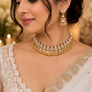 May include: A gold choker necklace featuring embedded white stones, small maroon beads, and dangling gold spheres, paired with matching gold drop earrings that include white stones, small white pearls, and dangling gold spheres.