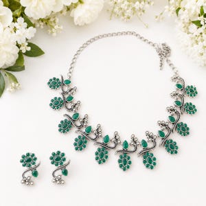 May include: A silver-toned necklace and matching pair of earrings featuring dark green, oval-shaped stones arranged in floral and teardrop patterns. The metalwork has an oxidized finish with intricate detailing, connected by a simple silver chain.