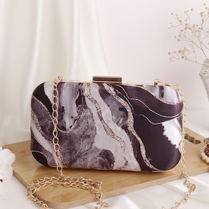 May include: A rectangular clutch with a gold chain strap. The clutch features a marble-like pattern in shades of black, gray, and white, with gold accents. The clasp and chain are gold-colored.