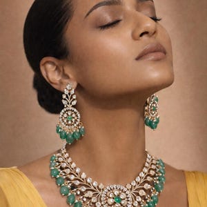 May include: A stunning jewelry set featuring a necklace and earrings. The necklace and earrings have a gold-tone metal base, adorned with clear crystals and vibrant green emerald beads. The necklace has a floral design, and the earrings are dangling.