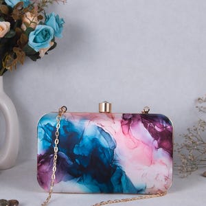 May include: Rectangular clutch with a gold chain strap. The clutch features a vibrant abstract design in shades of blue, pink, and purple. The clasp and frame are gold-toned, adding a touch of elegance to the accessory.