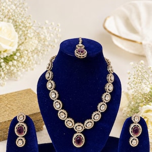May include: A jewelry set featuring a necklace, a pair of earrings, and a maang tikka. Each piece is crafted with oval-shaped deep purple stones surrounded by clear white crystals set in a metallic frame. The set is displayed on blue velvet stands.
