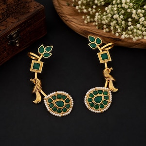 May include: A pair of gold-toned earrings featuring a peacock motif, accented with green stones and small white pearls. The design includes a leaf-shaped top section with three green stones and a square green stone, connected to a peacock figure.
