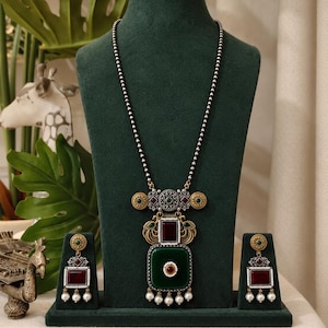 May include: A necklace and earring set featuring oxidized silver-tone metal with deep green and maroon square-shaped stones. The necklace has a long beaded chain and a large pendant with peacock motifs, while the earrings mirror the pendant design.