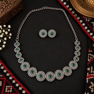 Può includere: Un set composto da collana e orecchini a lobo color argento, caratterizzato da medaglioni circolari in filigrana e maglie a forma di fiore, ciascuna con una pietra rotonda verde scuro. La collana presenta una catenina con chiusura a gancio.