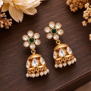 May include: A pair of gold-toned jhumka earrings featuring a flower-shaped stud with a central green stone surrounded by clear white stones. The bell-shaped drop is adorned with teardrop-shaped white stones and a row of small white pearl beads.