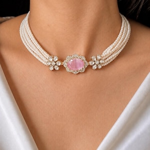May include: A multi-strand pearl choker necklace featuring a central oval pink gemstone set in a gold-toned frame, flanked by two floral-shaped accents encrusted with small clear stones, all connected to five rows of small white pearls.