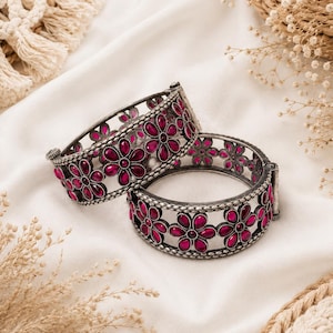 May include: Two hinged metal bangles featuring an oxidized silver finish and a repeating floral pattern. Each flower is composed of six teardrop-shaped, deep pink stones arranged around a central circular stone of the same vibrant color.