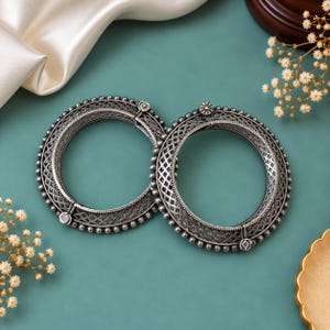 May include: A pair of circular silver-toned bangles featuring an intricate lattice pattern across the center band. The edges are lined with small spherical beads, and each bangle includes a hinged clasp mechanism with a small circular accent.