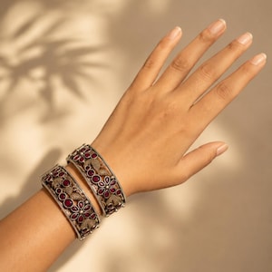 May include: Two wide metal bangles featuring an intricate oxidized silver finish with a textured, filigree-style pattern. Each bracelet is inlaid with deep red, teardrop-shaped stones arranged in floral motifs across the surface of the metal.