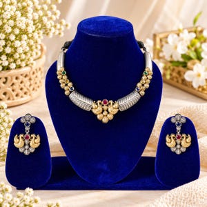 May include: A silver and gold-toned necklace and earring set featuring peacock motifs. The necklace has a central red stone, while the earrings feature matching red and blue stones. The metalwork includes textured silver segments and gold accents.