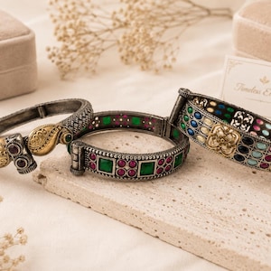 May include: Three hinged metal bangles featuring oxidized silver and gold-tone finishes. One bangle displays gold paisley motifs with dark red stones. Another features square green and round pink stones. The third has rows of multi-colored oval stones.