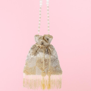 May include: A gold and silver beaded drawstring bag with a pearl and gold beaded handle. The bag has a gold tassel and a gold and silver beaded body. The background is a solid pink.