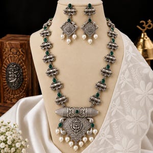 May include: A silver-toned necklace and earring set featuring oxidized metal detailing, green teardrop-shaped stones, and white pearl-like beads. The necklace includes a large central pendant with a face motif and matching drop earrings.