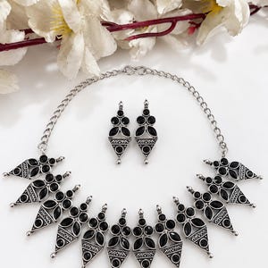 May include: A silver-toned necklace and matching earring set featuring triangular pendants with black stone inlays. The necklace consists of a chain with multiple dangling geometric charms, each accented with small black stones and intricate metalwork.