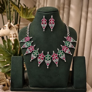 May include: A silver-toned necklace and earring set featuring triangular metal pendants inlaid with alternating pink and green teardrop-shaped stones. The necklace chain connects to a series of these geometric charms, while the matching earrings mirror the design with a single pendant each, showcasing intricate metalwork details.
