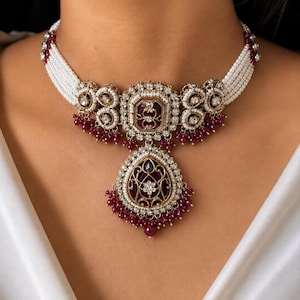 May include: A choker necklace featuring five strands of small white pearls, accented with gold-tone metal settings, clear crystals, and deep red stones. A large, pear-shaped pendant hangs from the center, adorned with red beads and crystals.