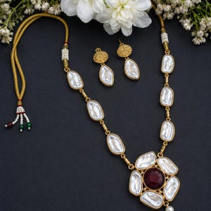 May include: A gold-toned necklace and earring set featuring oval-shaped white iridescent stones. The necklace includes a central floral-inspired pendant with a deep red circular stone at its center, accented by small white beads and a dangling pearl.