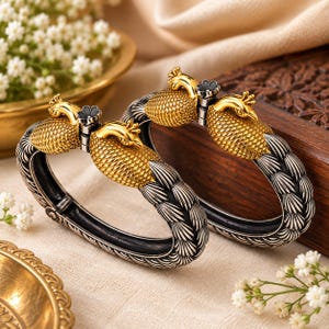 May include: A pair of hinged bracelets featuring gold-toned peacock motifs with textured bodies and silver-toned bands decorated with a repeating feather pattern. The peacock heads are detailed with small crests and dark accents on the floral elements.