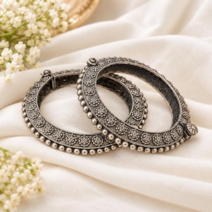 May include: Two silver-toned metal bangles featuring a repeating floral pattern along the centre band, bordered by rows of small spherical beads. Each bangle includes a hinged clasp mechanism with a circular decorative detail.
