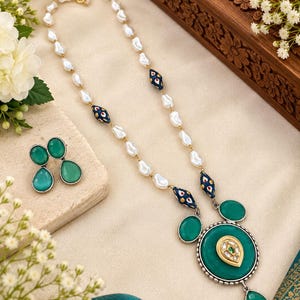 Puede incluir: Un collar con cuentas blancas irregulares, cuentas azul oscuro estampadas y un gran colgante circular con una piedra verde y un detalle central en forma de gota dorada. Pendientes verdes a juego acompañan al conjunto de collar.
