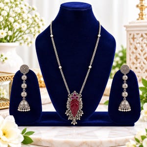 May include: A silver-toned necklace and earring set featuring oxidized metal detailing. The necklace has a central leaf-shaped pendant with a deep red stone and three small pink accents. The matching earrings are long, multi-tiered jhumka style.