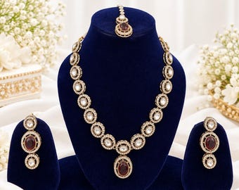 May include: A jewelry set featuring a necklace, two drop earrings, and a maang tikka. Each piece is crafted with gold-toned metal, circular frames, clear white stones, and a central deep red oval stone in a polished, ornate design.