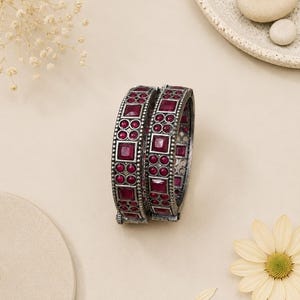 May include: Two hinged bangles featuring a dark oxidized silver-tone metal finish. Each bangle is inlaid with a repeating pattern of square and circular deep red stones, arranged in a geometric grid with beaded metal detailing along the edges.