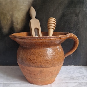 Puede incluir: Una jarra de cerámica marrón con asa, que contiene dos utensilios de cocina de madera. Uno es una espátula, el otro un cucharón de miel. La jarra tiene un borde ancho y una superficie texturizada, posiblemente vintage o hecha a mano. Altura aproximada 15 cm.