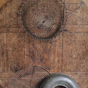 May include: An assortment of vintage kitchen tools, including a wire mesh strainer, a round cooling rack, and a metal donut-shaped mold. The items are arranged on a wooden surface with a floral painted background, evoking a rustic, antique aesthetic.