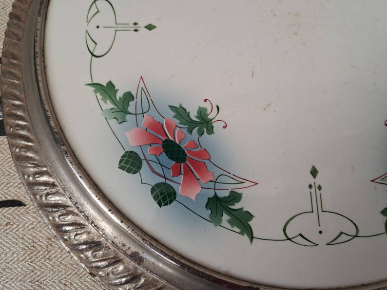 Op de afbeelding: Een wit keramiek bord met een zilveren rand en een bloemenmotief. Het ontwerp bevat roze bloemen, groene bladeren en blauwe accenten.