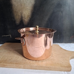 May include: A polished copper pot with a matching lid and a small side handle. The lid features a central brass knob. The pot has a cylindrical body with a smooth, reflective surface and a wide rim, showcasing a metallic finish throughout.