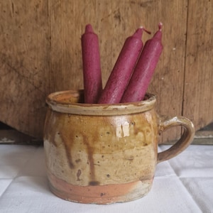 May include: A rustic, ceramic mug with a handle, filled with three deep magenta candles. The mug has a mottled, earthy color scheme with brown and orange tones. The background is a wooden surface.