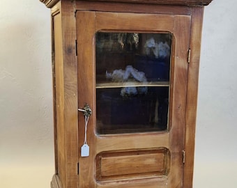 Op de afbeelding: Een rechthoekige houten wandkast met een enkele glazen deur, een kleine metalen grendel en een wit label. Twee langwerpige koperen sleutelplaten met sleutelgaten liggen horizontaal voor de onderkant van de kast.