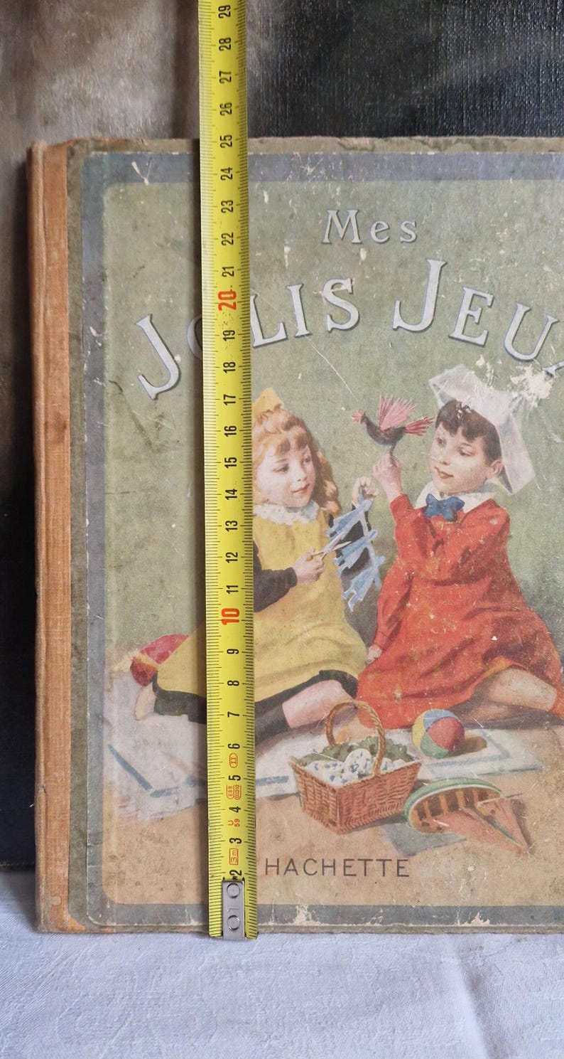 May include: Vintage children's book cover with the title "Mes Jolis Jeux." The cover features illustrations of two children playing with toys. The book measures approximately 10 inches tall. The publisher's name, HACHETTE, is printed at the bottom.