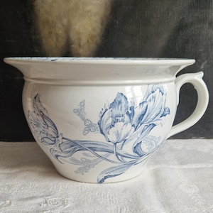 Puede incluir: Orinal de cerámica blanca con un diseño floral de tulipanes azules. El orinal tiene un borde ancho, un asa y un cuerpo redondeado. El patrón floral es detallado y cubre el exterior del orinal.