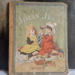 May include: Antique children's book titled "Mes Jolis Jeux" with a faded cover. The cover features two children in vintage clothing, with the publisher's name "Hachette" at the bottom. The book has a worn appearance, suggesting age and use.