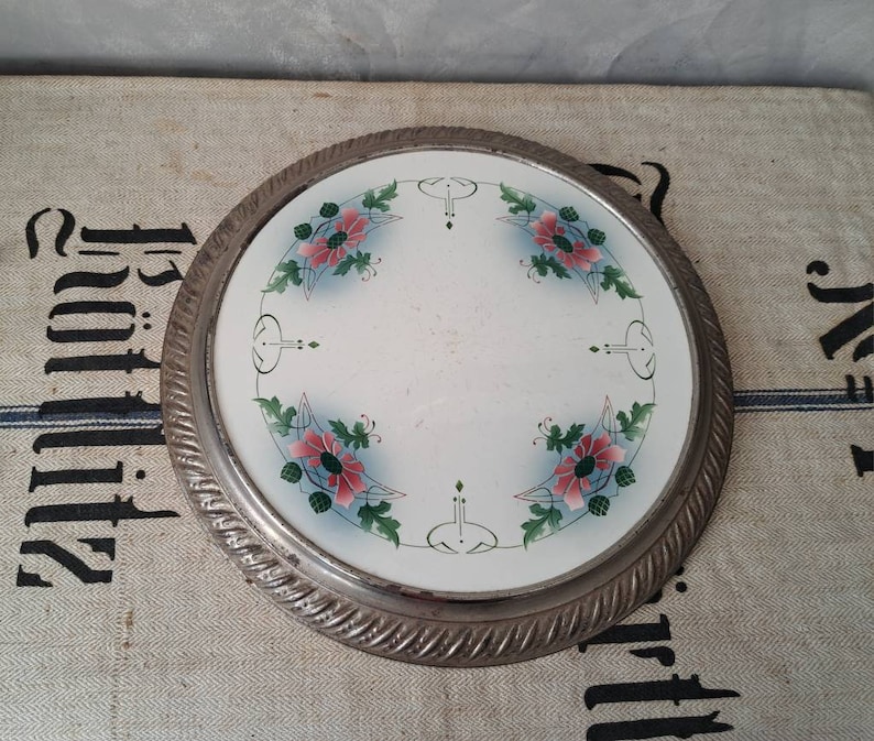 Op de afbeelding: Een ronde, zilveren randplaat met een wit centrum en een bloemenontwerp. De bloemen zijn roze en groen en hebben een gestileerd art nouveau-ontwerp.