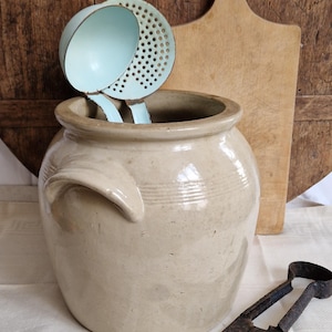 Op de afbeelding: Een beige keramische pot met een handvat, met twee lichtblauwe keukengerei. Een houten snijplank en antieke schaar staan op de achtergrond. De items zijn gerangschikt op een witte doek, met een rustieke houten achtergrond.